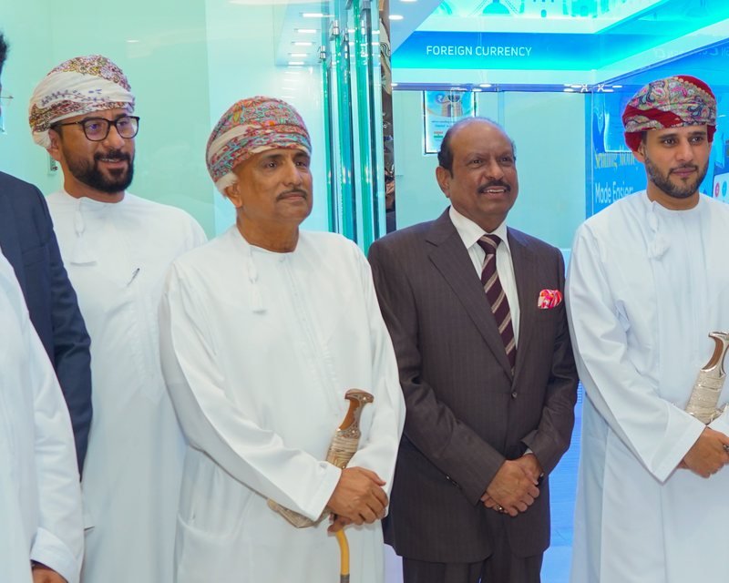 Two men standing side by side: one in a white Omani dress with a colourful embroidered cap and a cane, the other in a dark pinstriped suit with a striped tie and a red pocket square, in front of a sign that reads Foreign Currency